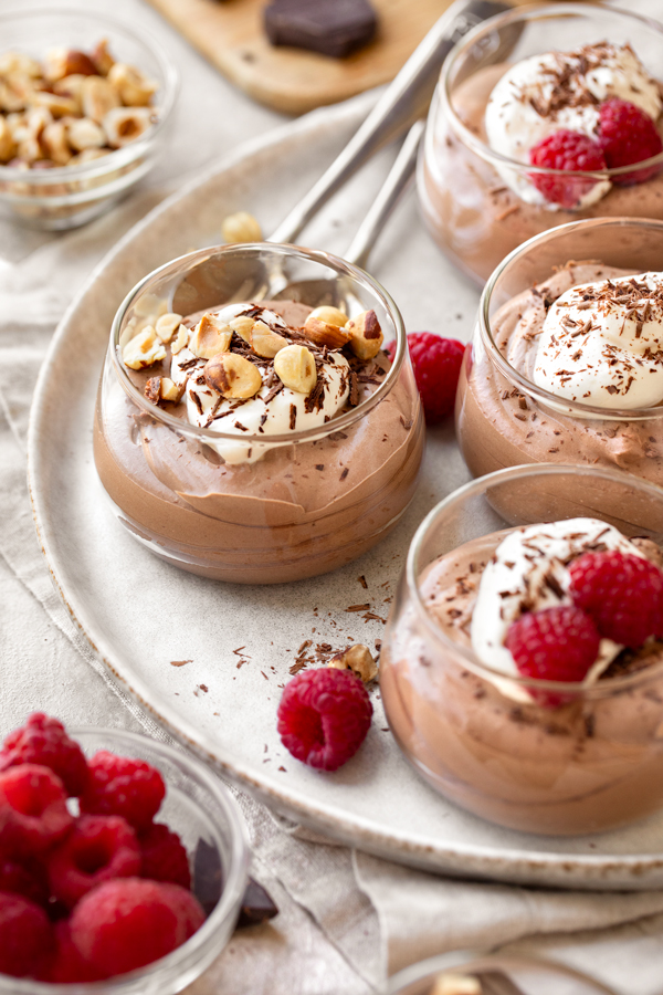 Four small glasses filled with Easy Chocolate Mousse, and topped with raspberries and/or toasted hazelnuts and chocolate shavings | thecozyapron.com