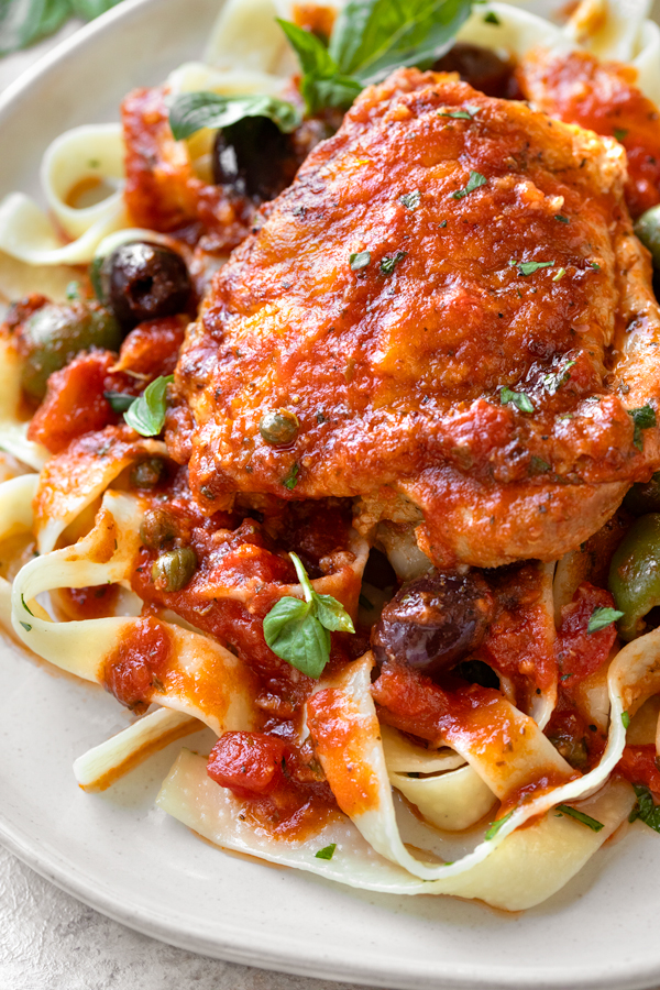 Closeup on a chicken thigh nestled in Puttanesca sauce over tagliatelle pasta | thecozyapron.com